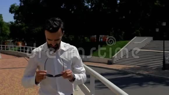 穿着白色衬衫留着胡须的时髦年轻人正走在一栋商业大楼附近戴上太阳镜4k4k视频视频的预览图