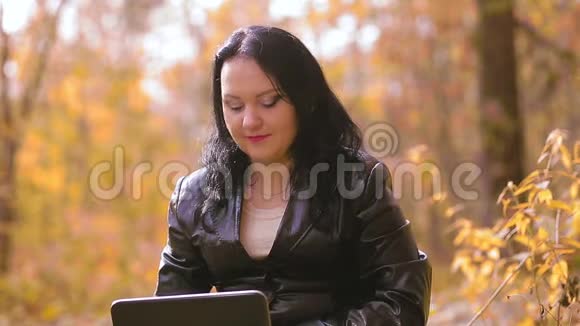 一个年轻的黑发女人正坐在一个阳光明媚的秋日公园里看着笔记本电脑屏幕微笑着视频的预览图