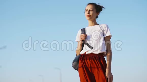 穿着白色t恤和红色裙子的年轻漂亮女人正站在蓝天的背景上看着视频的预览图