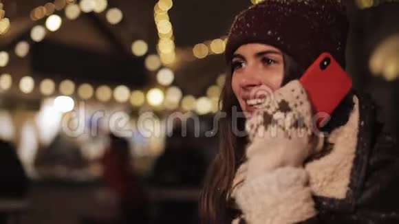 戴着冬帽和手套的漂亮女孩在智能手机上说话在下雪的时候笑着看着视频的预览图