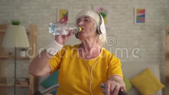 戴耳机的运动老妇人在运动自行车上进行有氧训练从一个瓶子里喝水视频的预览图