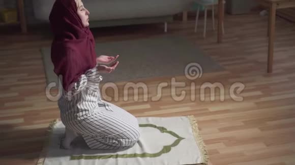 年轻的穆斯林妇女戴着传统的围巾坐在祈祷的位置上举手祈祷视频的预览图