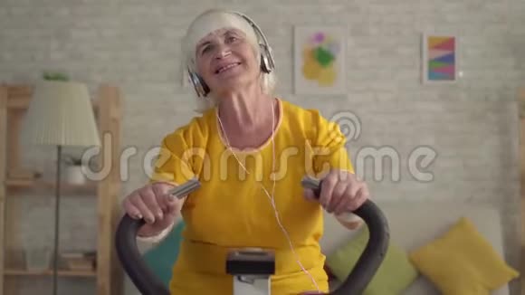 戴耳机的活跃老妇人在运动自行车上接受有氧训练视频的预览图