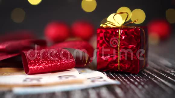一只手在红色礼品盒和欧元钞票旁边放着一颗装饰性的粉红色心明亮闪亮的蝴蝶结特写和圣诞球视频的预览图