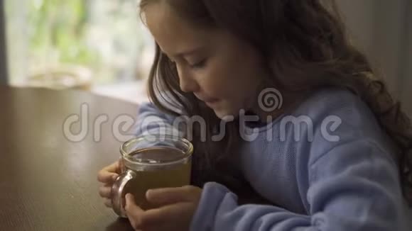 白种人的小女孩长发卷曲吹在一杯洋甘菊茶上一个穿着蓝色衣服的可爱的黑发孩子视频的预览图