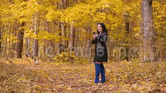秋天公园里一个穿着蓝色靴子的黑发年轻女子站在一棵树旁打电话视频的预览图