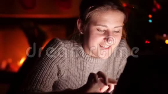 4k镜头微笑的年轻女子躺在壁炉旁用数码平板电脑晚上喝热茶视频的预览图