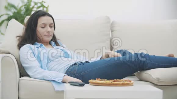漂亮的女人躺在家里的沙发上一边吃披萨一边看电视4K视频的预览图