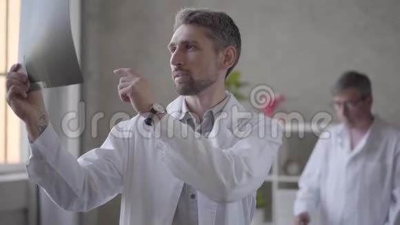 两名男医生在现代诊所的办公室检查x光穿白色长袍的年轻人给他的年长者看x光视频的预览图
