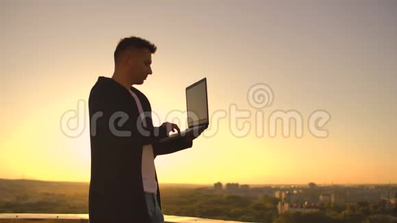 日落时分一位男性股票经纪人自由职业者站在屋顶上手提电脑和键盘上的键盘视频的预览图
