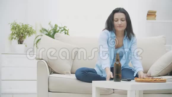 一个年轻的女人坐在沙发上吃披萨喝啤酒她正在打开电视好吃好吃4K视频的预览图