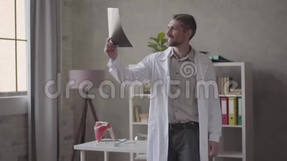 英俊的积极的成功的满意的男人穿着白色长袍手里拿着x光在现代诊所跳舞医生视频的预览图