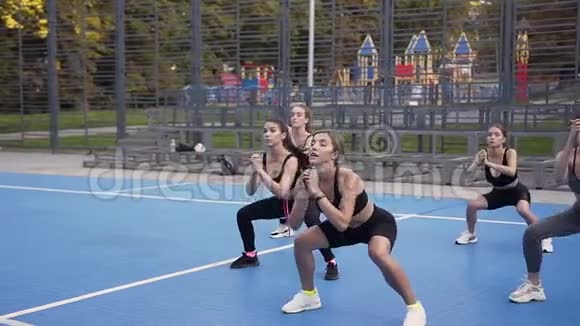 穿着运动服的年轻苗条的女孩子白天在绿树环绕的运动场上做蹲式运动视频的预览图