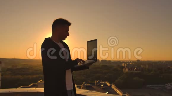 日落时分一位男性股票经纪人自由职业者站在屋顶上手提电脑和键盘上的键盘视频的预览图