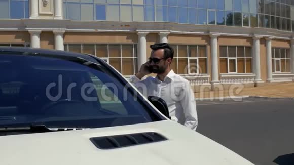 一个留着胡须的年轻男子穿着白色衬衫戴着深色眼镜站在他的白色汽车附近打电话4k4k视频视频的预览图