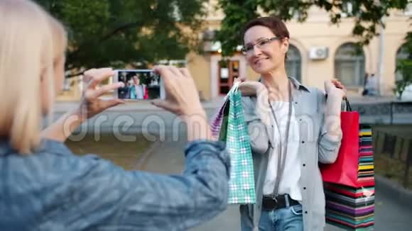 有魅力的女士带着购物袋在户外摆摆智能手机摄像头视频的预览图