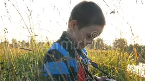 日落时分小男孩聚精会神地看着草地上的摄像机屏幕视频的预览图