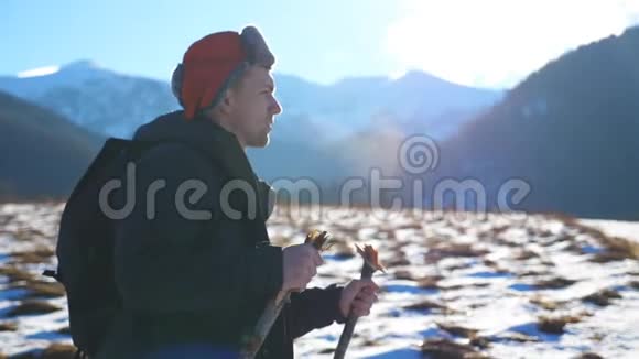 山背景下年轻的徒步旅行者在雪地小径上用棍棒行走在野外的侧面观看运动男视频的预览图