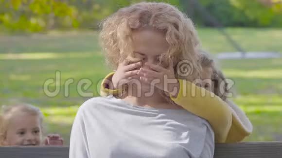 秋天公园里坐在长凳上的年轻白种人卷曲的金发女人的特写肖像她的两个小可爱视频的预览图