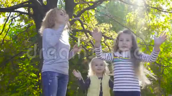 年轻的金发女人呆在阳光明媚的公园里吹肥皂泡她的两个小漂亮的女儿视频的预览图