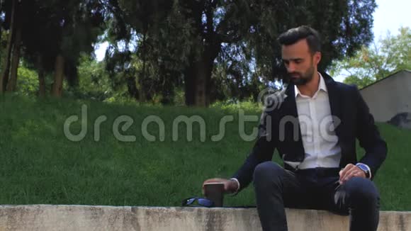 迷人的商人留着一身经典西装的胡须坐在草地背景下的公园里看着电话喝着茶或视频的预览图
