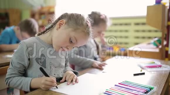 女孩双胞胎姐妹在教室的一张桌子上用毛毡笔在纸上画画学龄前儿童在学校接受艺术教育视频的预览图