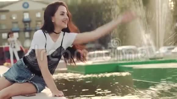 穿着牛仔短裤的年轻女人在城市喷泉里撒娇地在水中嬉戏溅起水花视频的预览图