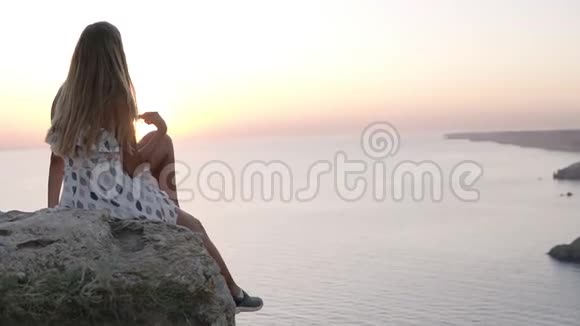 美丽的少女坐在海边的岩石上欣赏着夕阳无忧无虑地挥动着她的腿视频的预览图