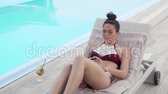 美丽的女人在泳池边休息时用智能手机自拍视频的预览图