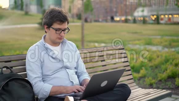 年轻人在公园里的笔记本电脑上打字坐在公园背景模糊的长凳上视频的预览图