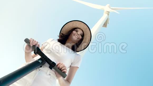 一个女孩戴着草帽和电动滑板车在风力发电机背景下的肖像视频的预览图