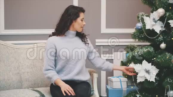 漂亮的年轻女人坐在圣诞树附近的沙发上享受着节日的精神视频的预览图