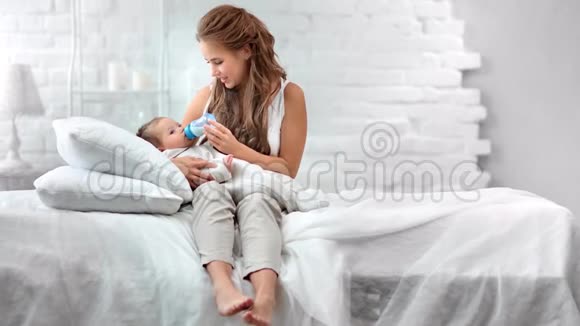 赤脚微笑的女人在白色房间里躺在床上享受着从奶瓶中喂养新生婴儿的乐趣视频的预览图