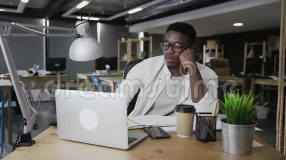 疲惫的非洲男人在笔记本电脑上工作等待着一天的工作结束视频的预览图