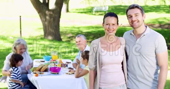 母亲和父亲在镜头前微笑他们的家人在背后吃午餐视频的预览图