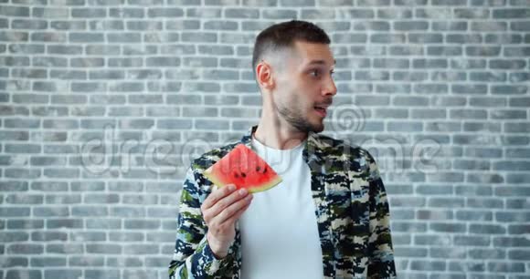 一幅英俊男子吃着新鲜西瓜在砖背景上微笑的画像视频的预览图