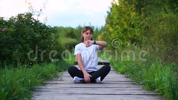 苗条的运动女孩坐在木桥上摆姿势看着手表视频的预览图