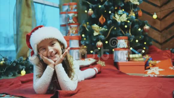 圣诞老人的孩子躺在圣诞树下的红毯子上把她的头从一边移动到另一边把手放在上面视频的预览图
