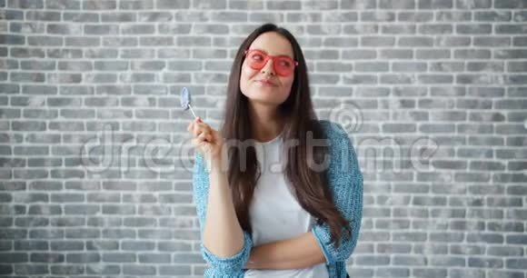 戴着太阳镜的快乐女孩拿着棒棒糖在砖墙背景下微笑视频的预览图