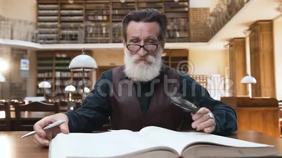 坐在图书馆桌旁的一位长着漂亮胡子的老人摘下眼镜在相机上摆姿势合上书视频的预览图