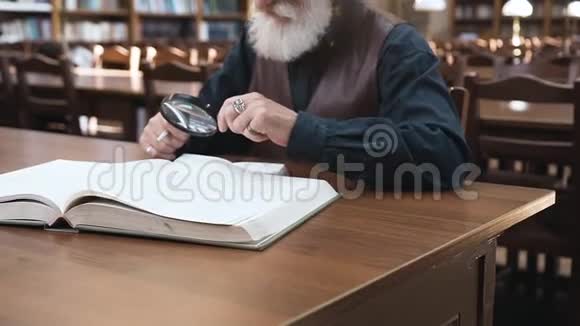 坐在图书馆里用放大镜与书合作的戴着眼镜的长胡子老人的侧视图视频的预览图