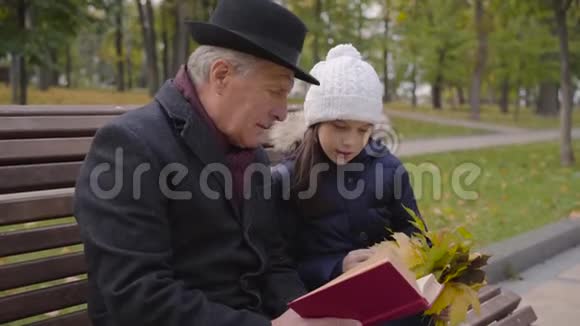 一个穿着经典服装的白种人成年男子和孙女坐在长凳上读着一本红色封面的书视频的预览图