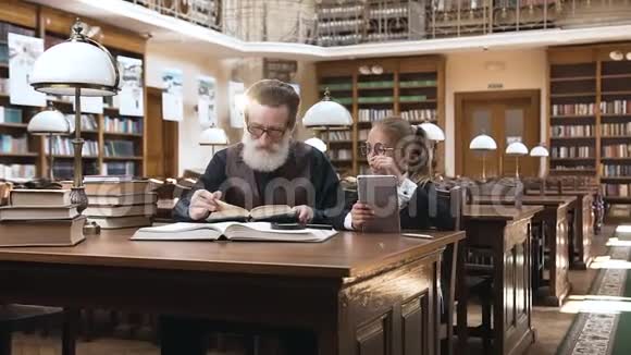 聪明的老胡子爷爷戴着眼镜在图书馆里读他10岁的孙女有趣的书视频的预览图