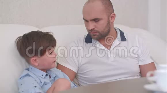 微笑的父亲和坐在白色沙发上喝茶的儿子说话视频的预览图
