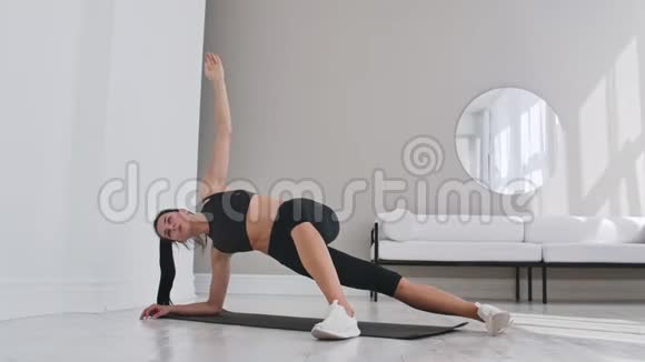 年轻性感的黑发女人穿着运动服在白色的内部做侧板运动在家里倾斜慢动作视频的预览图