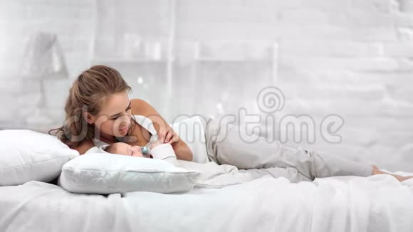 可爱的微笑妈妈躺在舒适的白色床上逗乐可爱的宝宝有积极的情绪视频的预览图