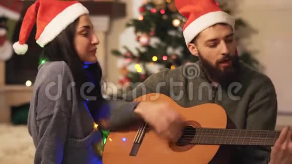 圣诞老人戴着帽子在家唱歌的幸福夫妇的肖像弹吉他的人脖子上戴着花环的女人视频的预览图