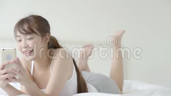 美丽的肖像年轻的亚洲女人微笑着躺着看智能手机阅读社交网络与享受床上卧室视频的预览图