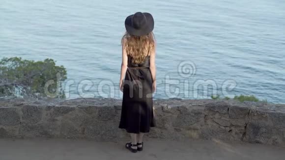 穿着黑色丝绸连衣裙和黑色帽子的长发女孩呆在蔚蓝海湾的小径上晴天碧海松林视频的预览图