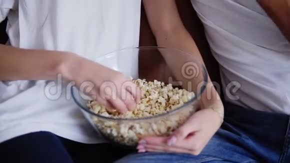 一对优雅的夫妇的特写他的手拿着一个大透明的碗在家吃爆米花一对穿着蓝色牛仔裤视频的预览图